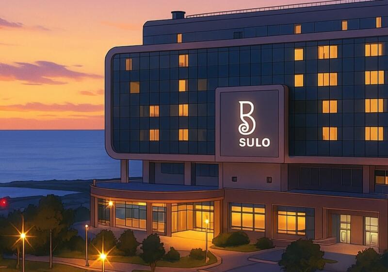 A modern building with a sign that reads "SULO", featuring a stylized "B", stands near a body of water under a colorful sunset sky.