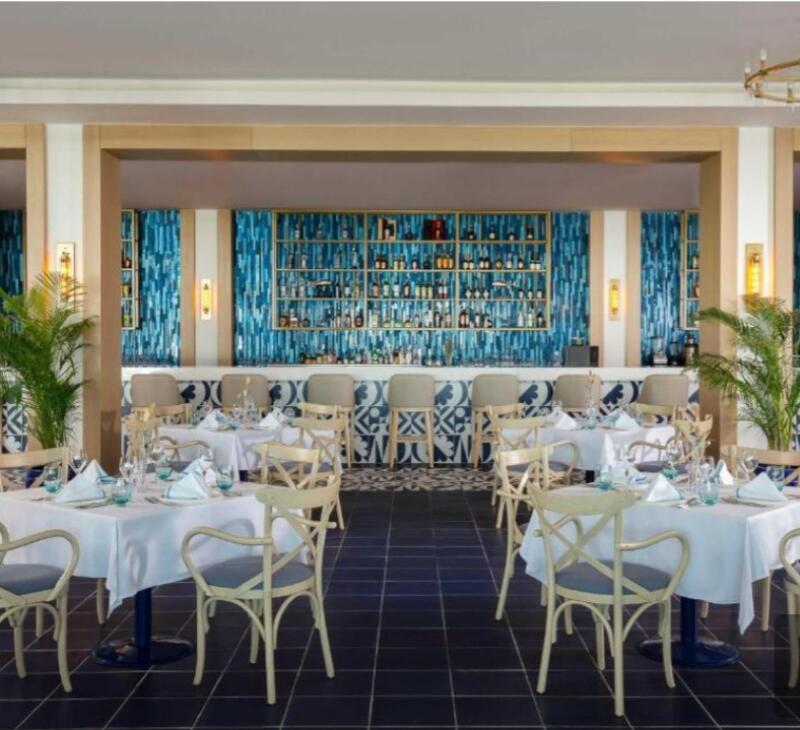 The interior of a restaurant features tables with white linens and light-colored chairs, a bar with blue patterned stools, and shelves stocked with bottles.