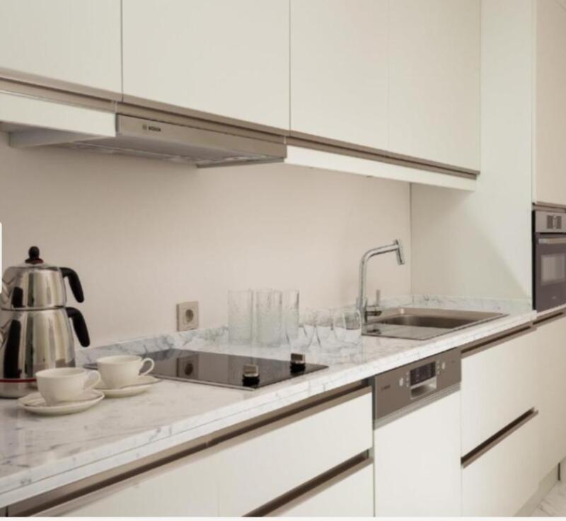 A kitchen with white cabinets and a marble countertop features a tea kettle, cups, glasses, a sink, and a stovetop.