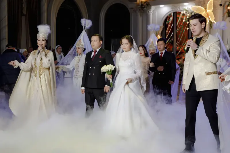 A wedding procession moves through a cloud of fog, led by a woman in traditional attire singing into a microphone, followed by the bride and groom, and other traditionally dressed individuals.