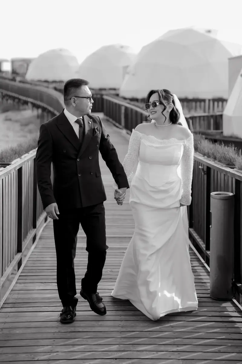 A black and white photo of a couple in wedding attire walking hand-in-hand on a wooden bridge, with white dome-shaped structures visible in the background. The bride wears sunglasses and a pearl necklace.
