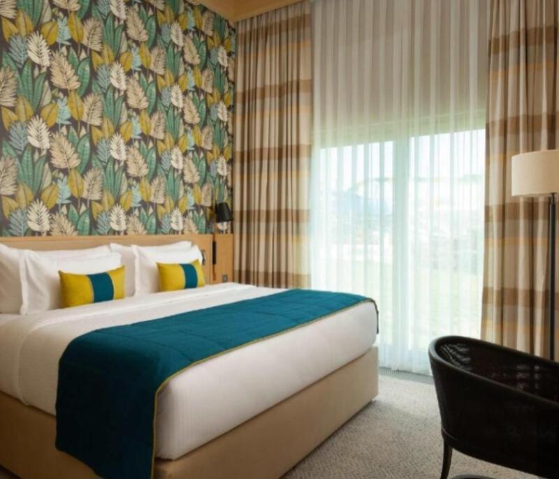 A bedroom features a bed with white linens, a blue runner, and yellow and blue pillows, complemented by leaf-patterned wallpaper and striped curtains.