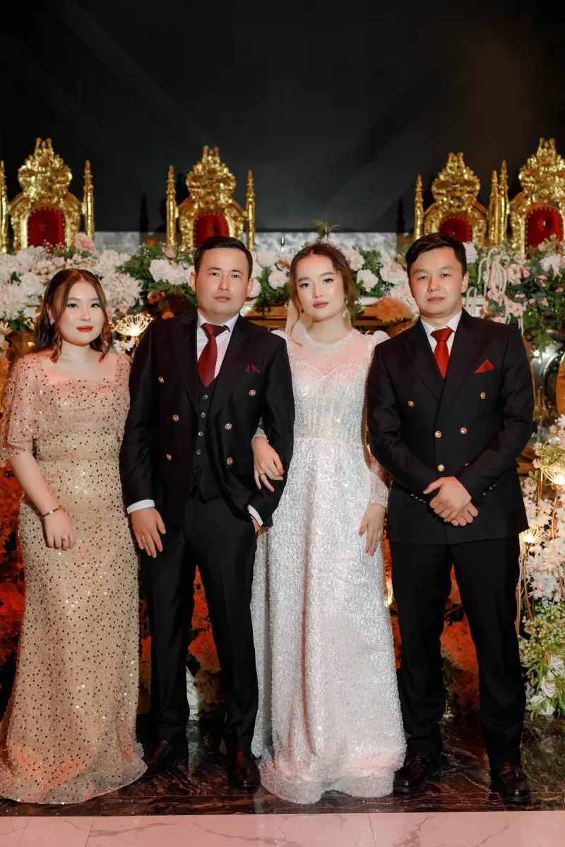 A group of four people stands in front of a floral and golden throne backdrop; the bride is wearing a white dress, one woman wears a gold sequined dress, and two men are in black suits with red ties.
