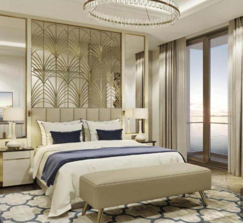 The bedroom features a large bed with white linens, navy pillows, and a blue throw, complemented by a beige bench at the foot, and a decorative gold headboard. The room also includes side tables with lamps, a crystal chandelier, floor-to-ceiling windows with curtains, and a patterned rug.