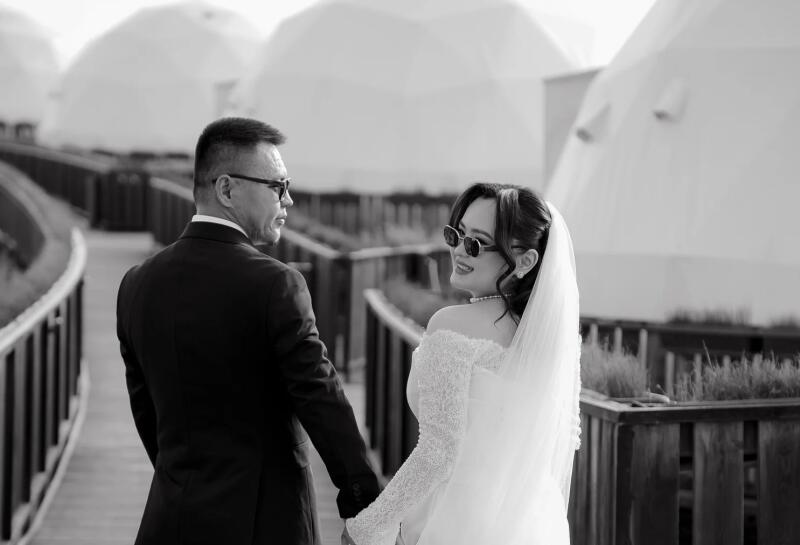 A black and white photo of a groom and bride walking hand-in-hand on a boardwalk, with the bride looking back and smiling while wearing sunglasses and a veil. White dome structures are visible in the background.