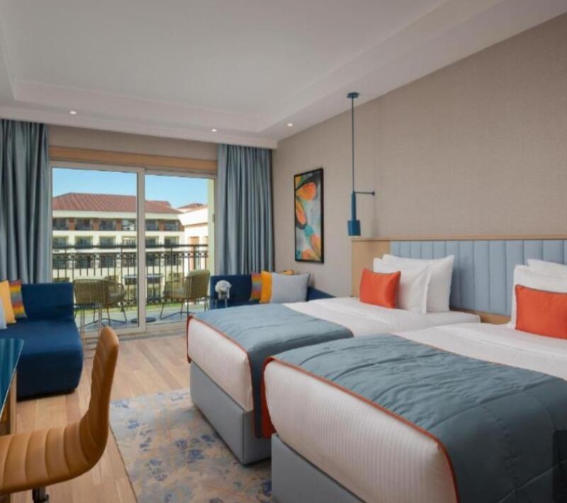 A hotel room features two twin beds with blue comforters, orange pillows, and a blue headboard. There is also a seating area with a blue couch and a balcony with chairs.