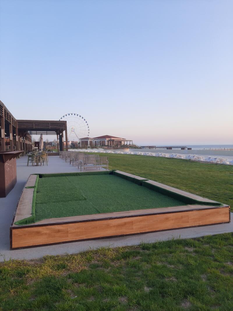 The image shows a bocce ball court with artificial grass, surrounded by a wooden frame, in an outdoor setting near a beach with a Ferris wheel in the background.