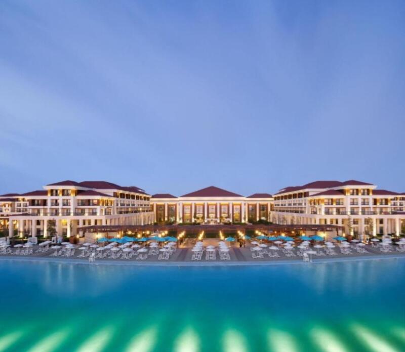 The image shows a large hotel complex with a red roof near a body of water, possibly a sea or a large pool. The hotel has multiple floors and is surrounded by beach umbrellas and chairs.