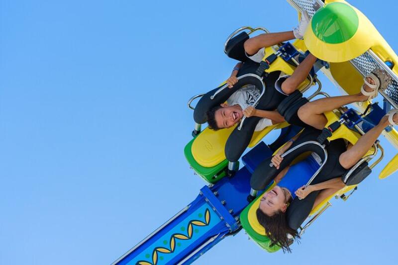 Two people are on an amusement park ride against a clear blue sky, strapped into seats on a colorful rotating apparatus.