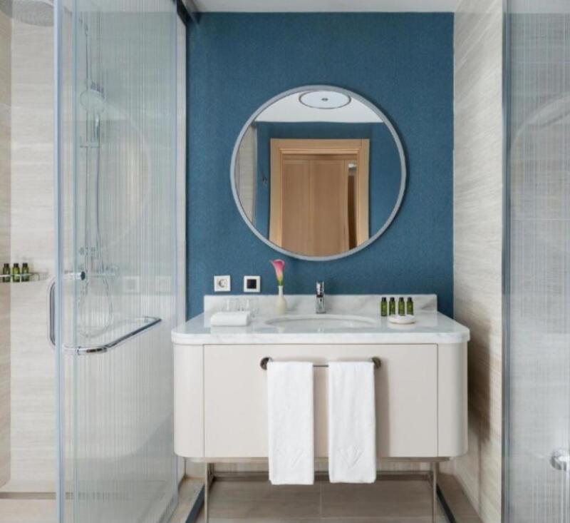 A bathroom features a glass shower stall on the left, a white sink and vanity with two white towels hanging from it, and a round mirror on a blue wall.