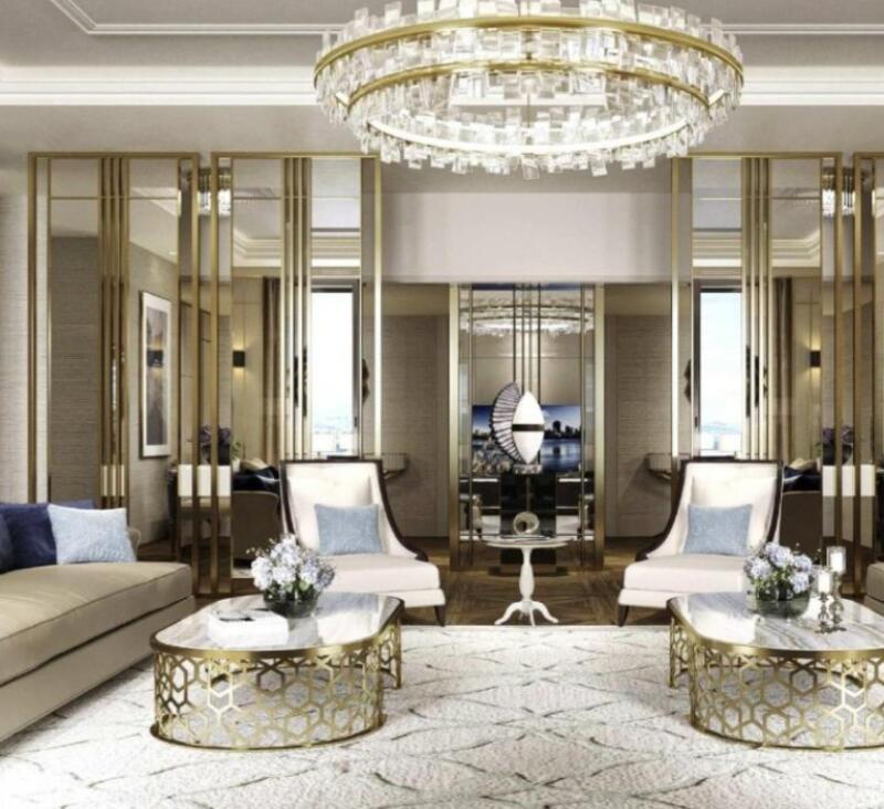 The image shows a luxurious living room with a large crystal chandelier, two chairs with light blue pillows, a beige sofa with blue pillows, two coffee tables with gold geometric bases, and mirrored wall panels.