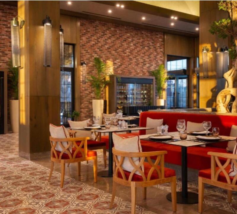 The interior of a restaurant features tables set with glassware and tableware, surrounded by wooden chairs with red cushions and a red upholstered banquette. A brick wall and potted plants add to the ambiance.