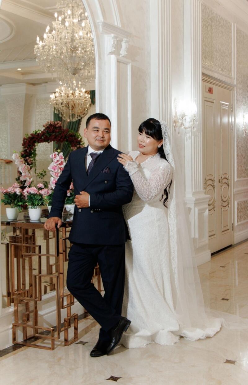 A man in a dark blue suit and a woman in a white wedding dress are posing in an ornate hall with a chandelier and floral decorations.