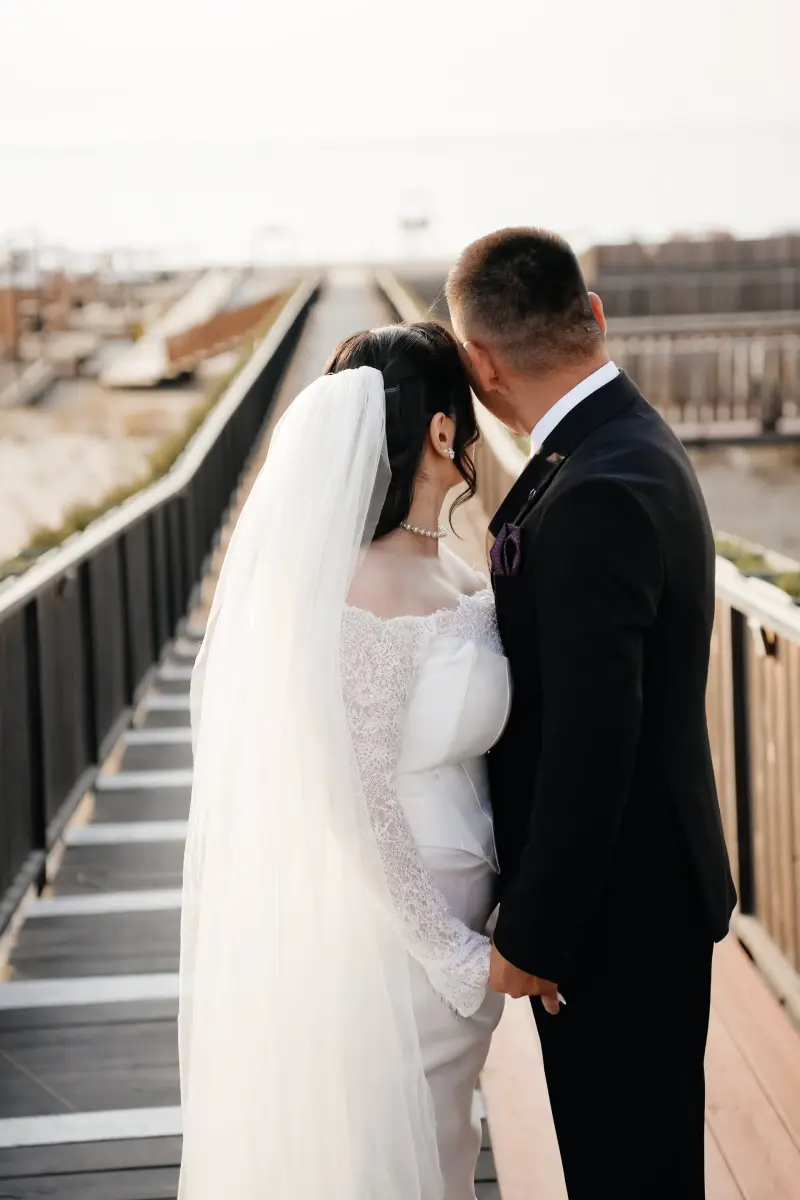 A bride in a white lace wedding dress and veil stands hand-in-hand with a groom in a black suit, viewed from behind on a wooden walkway, looking into the distance.