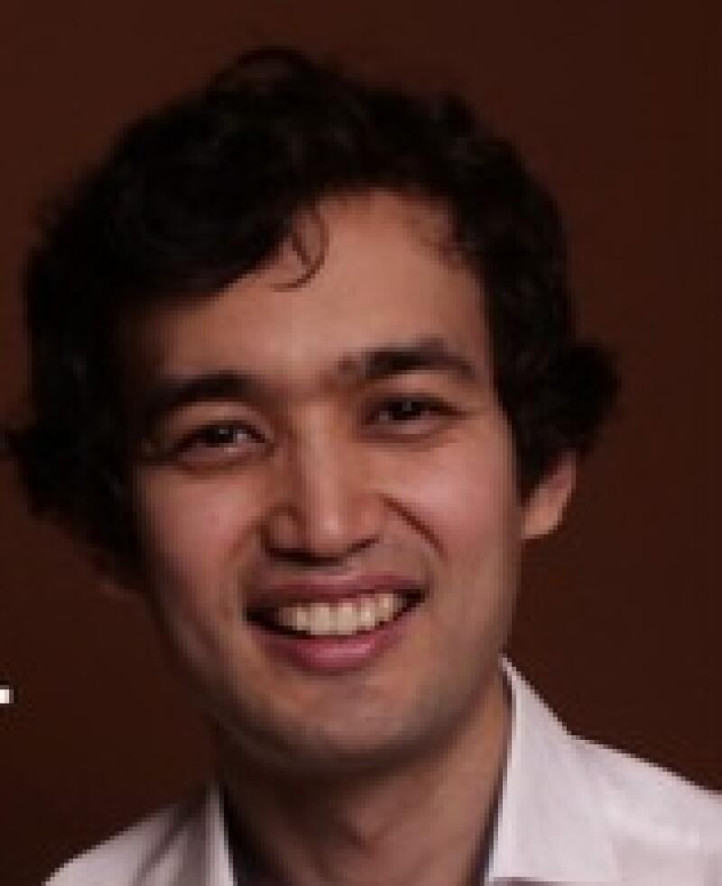 A smiling man with dark, curly hair and a white collared shirt is set against a brown backdrop.