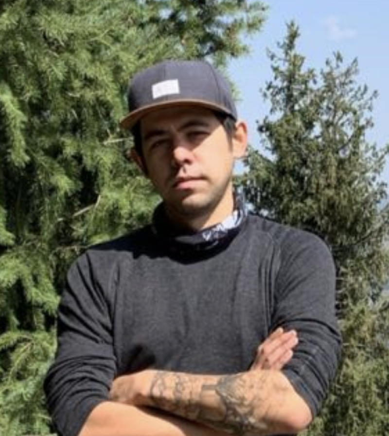 A person with crossed arms, wearing a black long-sleeve shirt and a baseball cap, stands in front of green trees; tattoos are visible on their left forearm.