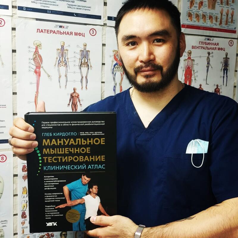 A man in a blue medical scrub is holding a book titled 'Manual Muscle Testing: A Clinical Atlas' by Gleb Kirdoglo, with anatomical charts visible in the background.