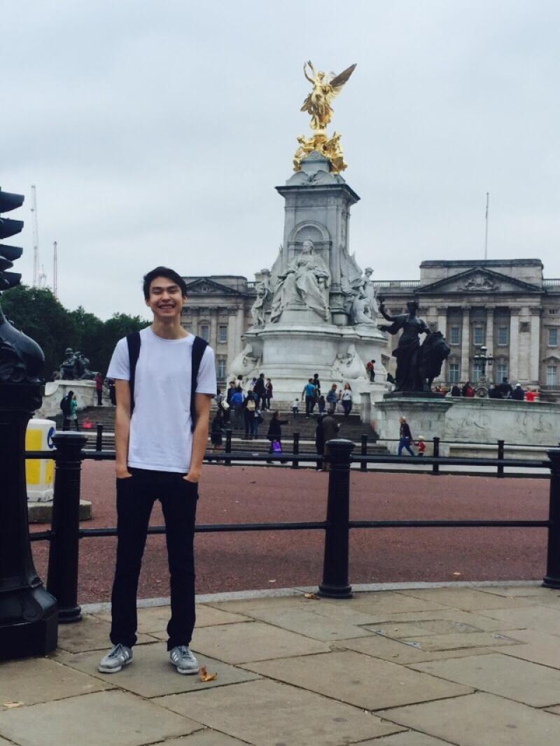 A young man wearing a white t-shirt and black pants stands in front of the Victoria Memorial at Buckingham Palace.