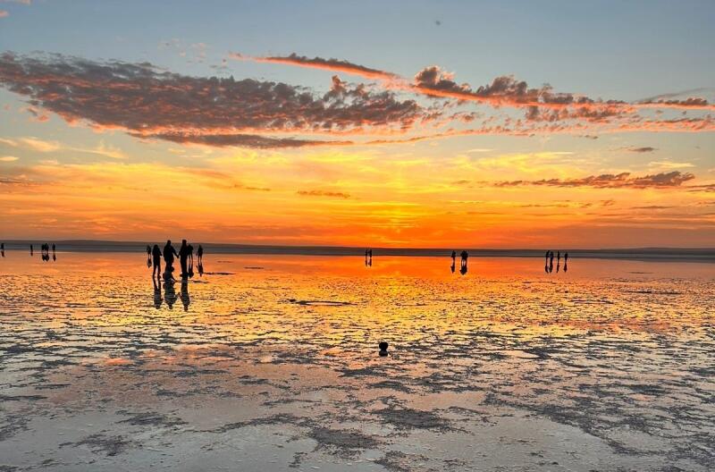 The photograph depicts a sunset scene over a body of water, with several people silhouetted against the vibrant orange and yellow sky.