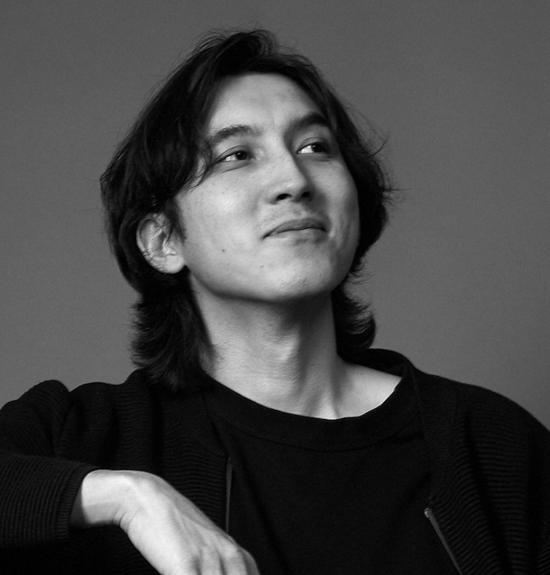 A black and white portrait of a man with long hair, wearing a dark shirt and jacket.