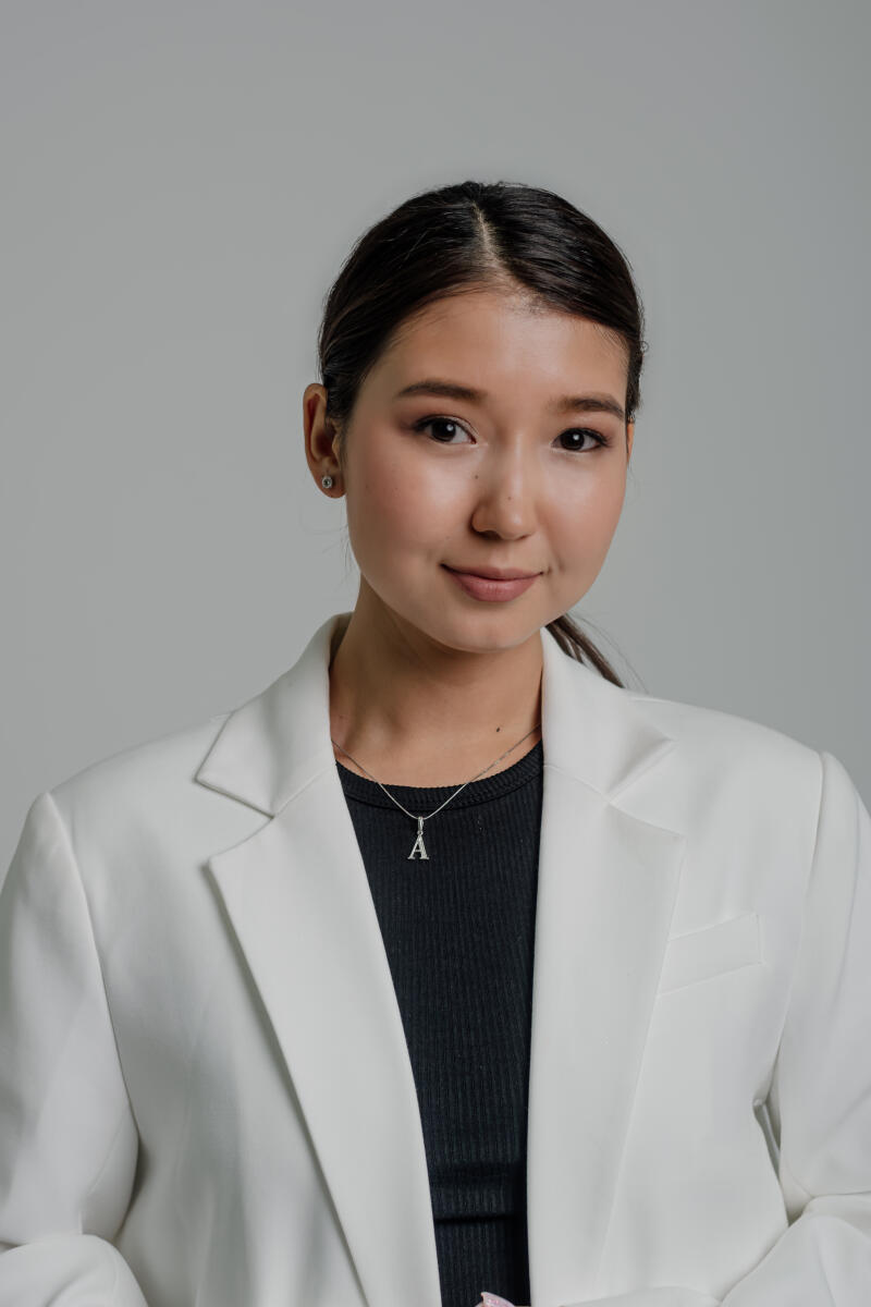 A woman with dark hair pulled back, wearing a white blazer over a black top and a necklace with a pendant in the shape of the letter "A".