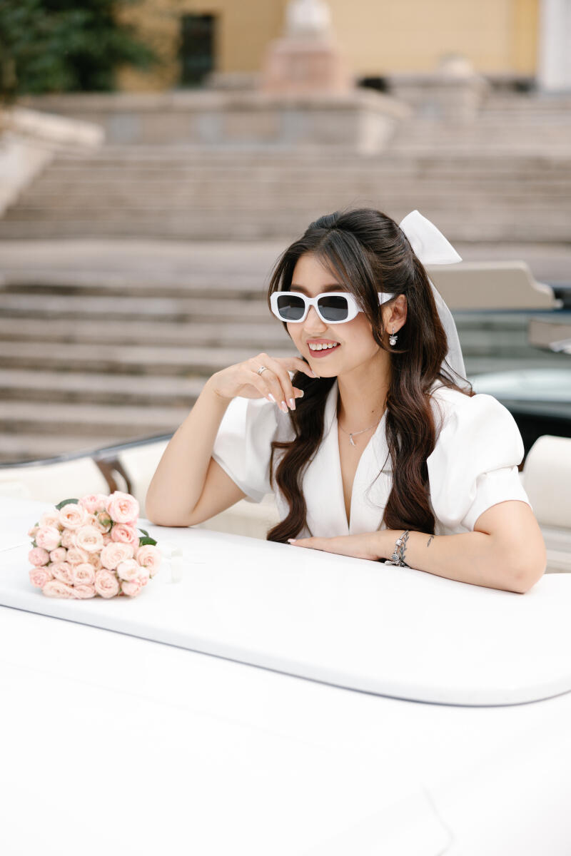 A smiling woman with long brown hair, wearing white sunglasses and a white dress, sits in a car with a bouquet of light pink roses on the hood.