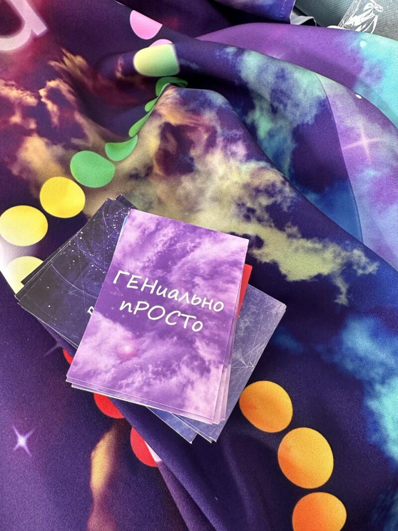 A stack of cards with the text "ГЕНИАЛЬНО просто" (GENIALNO prosto) lies on a colorful fabric with a space theme, featuring nebulae, stars, and colored dots.
