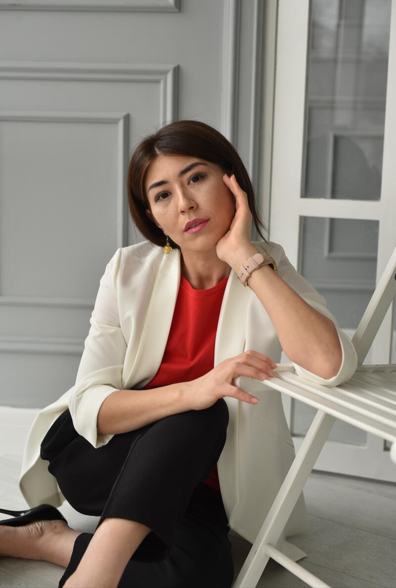 A woman wearing a white blazer and black pants sits on the floor, leaning against a white chair.