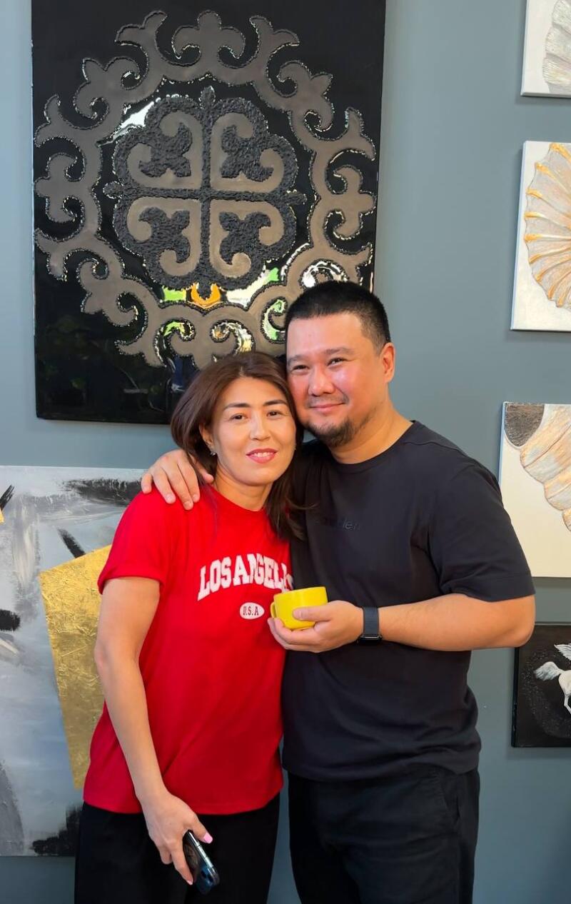 A woman in a red "Los Angeles" T-shirt and a man in a black T-shirt are posing together in front of artwork, with the man's arm around the woman and holding a small yellow cup.