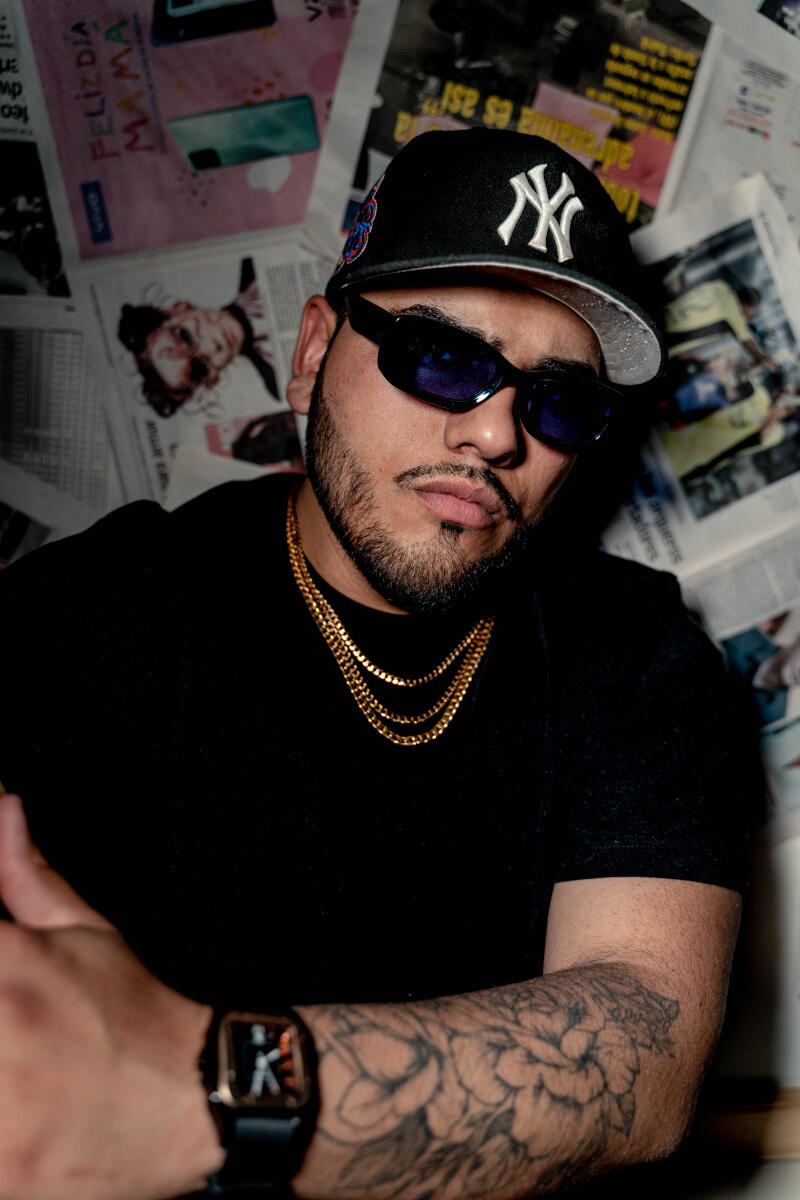 A man with a beard, wearing a black shirt, gold chains, a black New York Yankees baseball cap, and sunglasses, poses with a tattoo on his arm in front of a newspaper-covered wall.