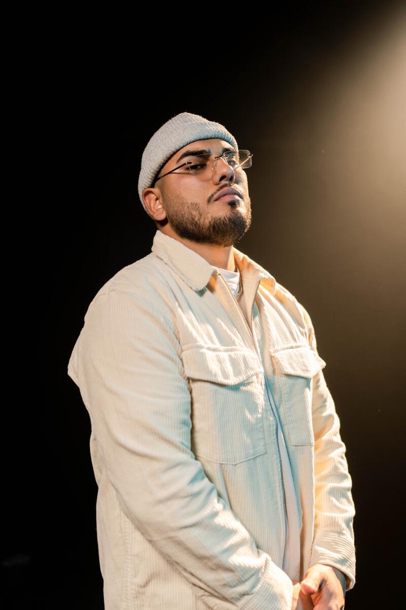 A man with a beard, wearing glasses and a beanie, is posing in a light-colored corduroy jacket against a dark background.