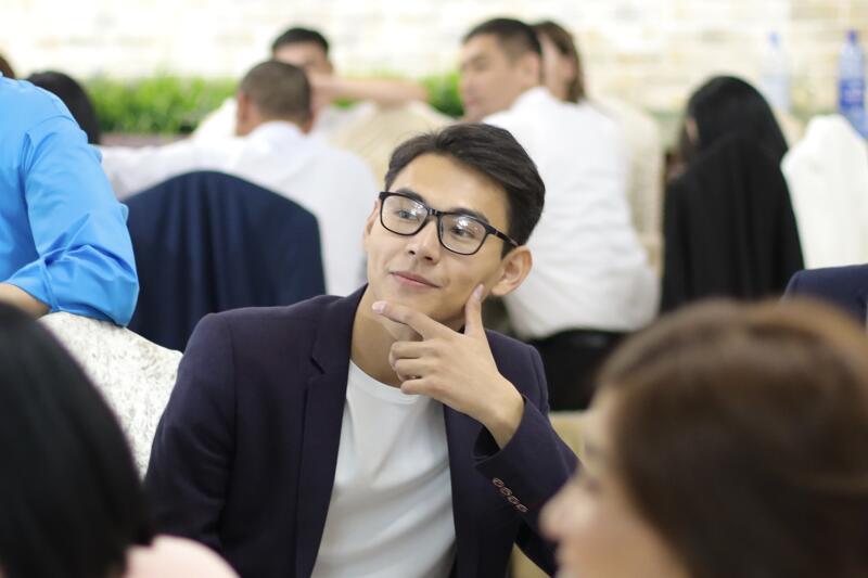 A man wearing glasses and a blazer rests his chin on his hand while looking off to the side in a crowded room.