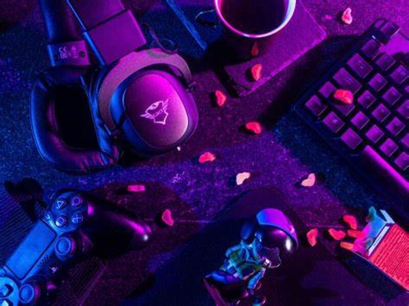 The photo features a gaming setup with a headset, controller, keyboard, a cup of coffee, and a small statue, all illuminated in blue and purple lighting with scattered heart-shaped candies.