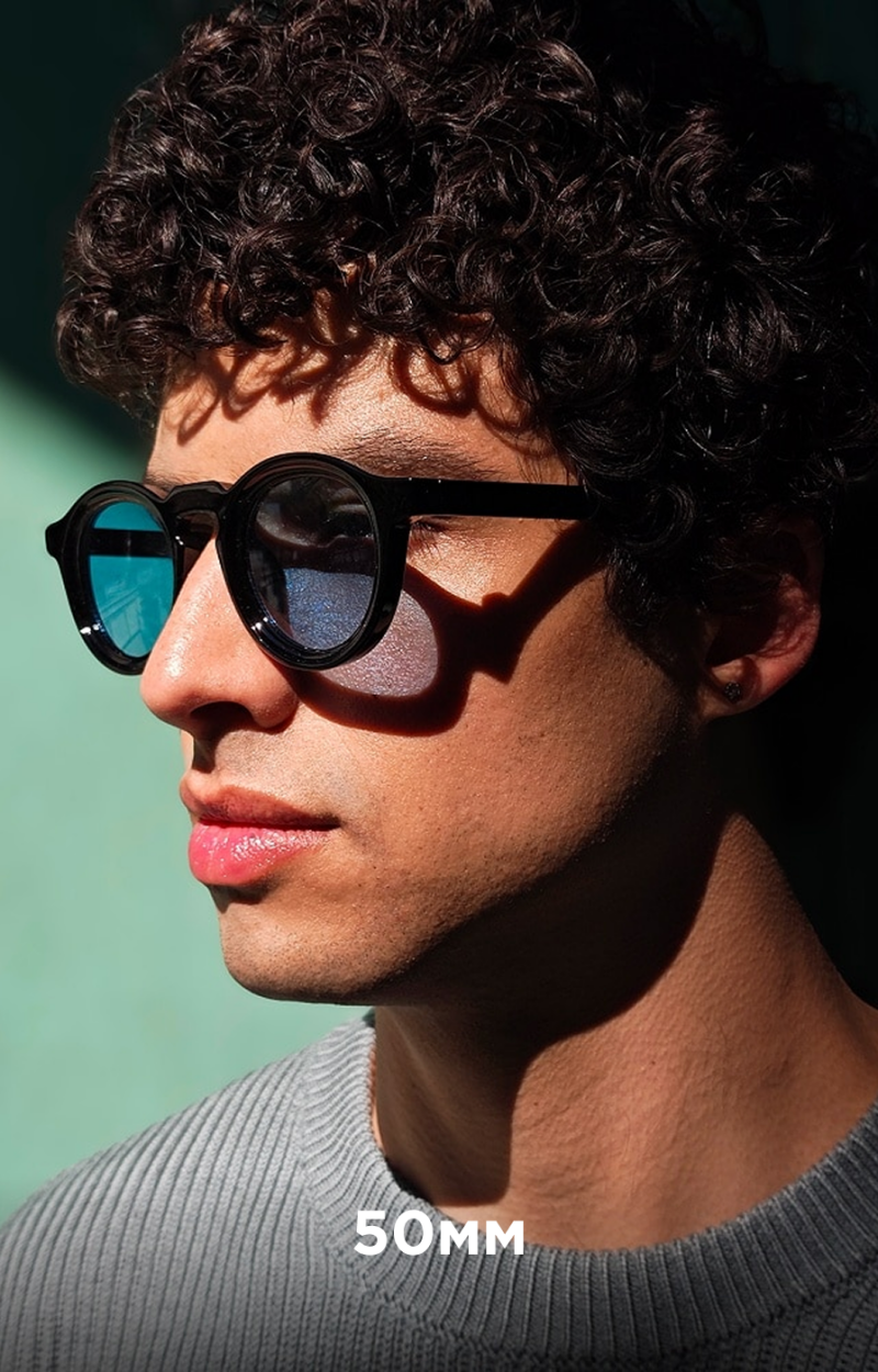 A person with curly hair wears black sunglasses and a gray sweater, with the text "50MM" visible on the sweater.