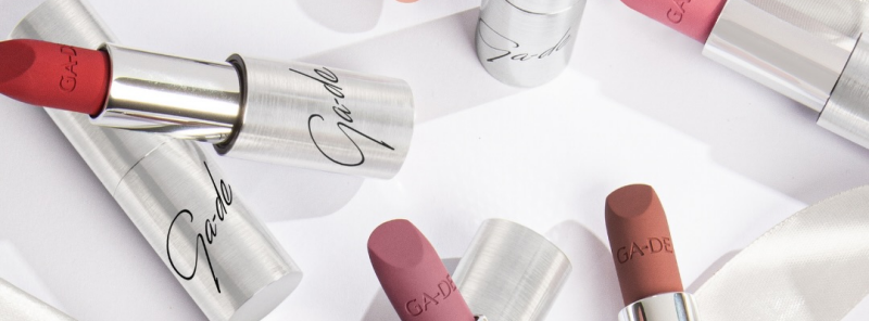 The image shows several lipsticks of different colors with silver tubes labeled 'Ga-de' scattered on a white surface.
