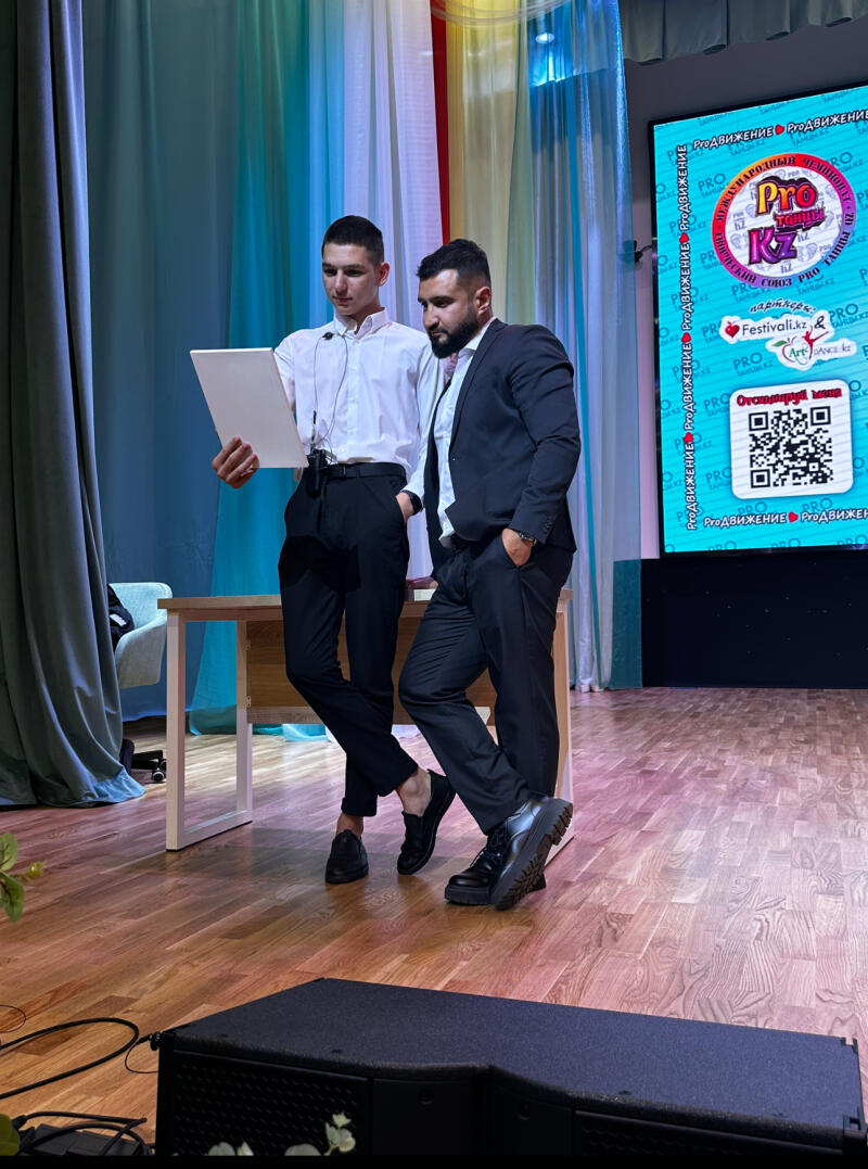 Two men are standing on a stage in front of a screen that displays the word 'PRO' and a QR code; one man is holding a white board, and the other is leaning against a table, wearing a suit.