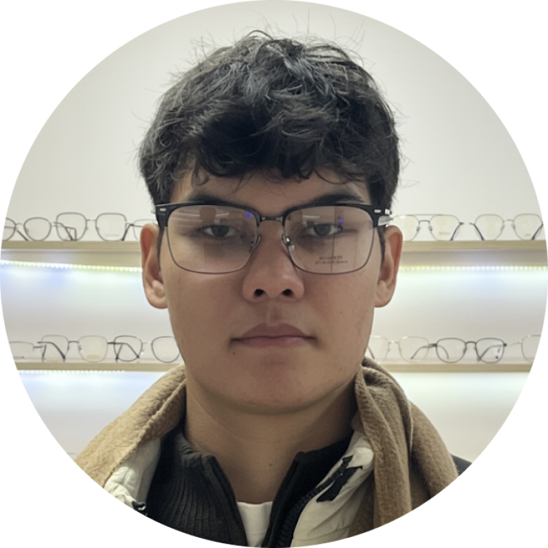 A close-up shot of a person with dark hair and glasses, wearing a jacket with a scarf around their neck, with a display of eyeglasses in the background.