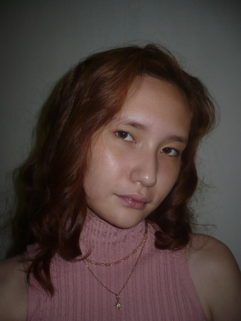 A woman with wavy auburn hair and fair skin wears a light pink top and two gold necklaces, one with a star pendant.