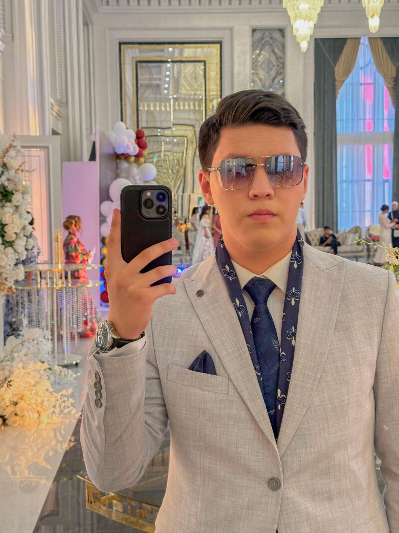 A man takes a selfie in a light gray suit, blue tie, and sunglasses, with a watch on his left wrist. The background shows a fancy room with decorations, balloons, and other people.