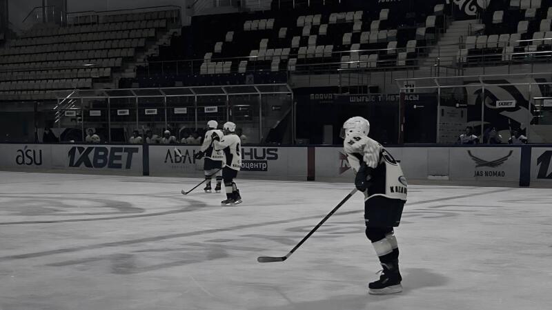 The black and white image shows hockey players on the ice during a game, with logos such as "1XBET" and "JAS-NOMAD" visible on the rink's boards.