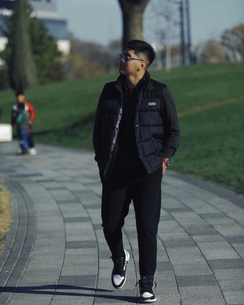 A young man in a black vest and pants walks on a brick path, looking upwards and wearing glasses.
