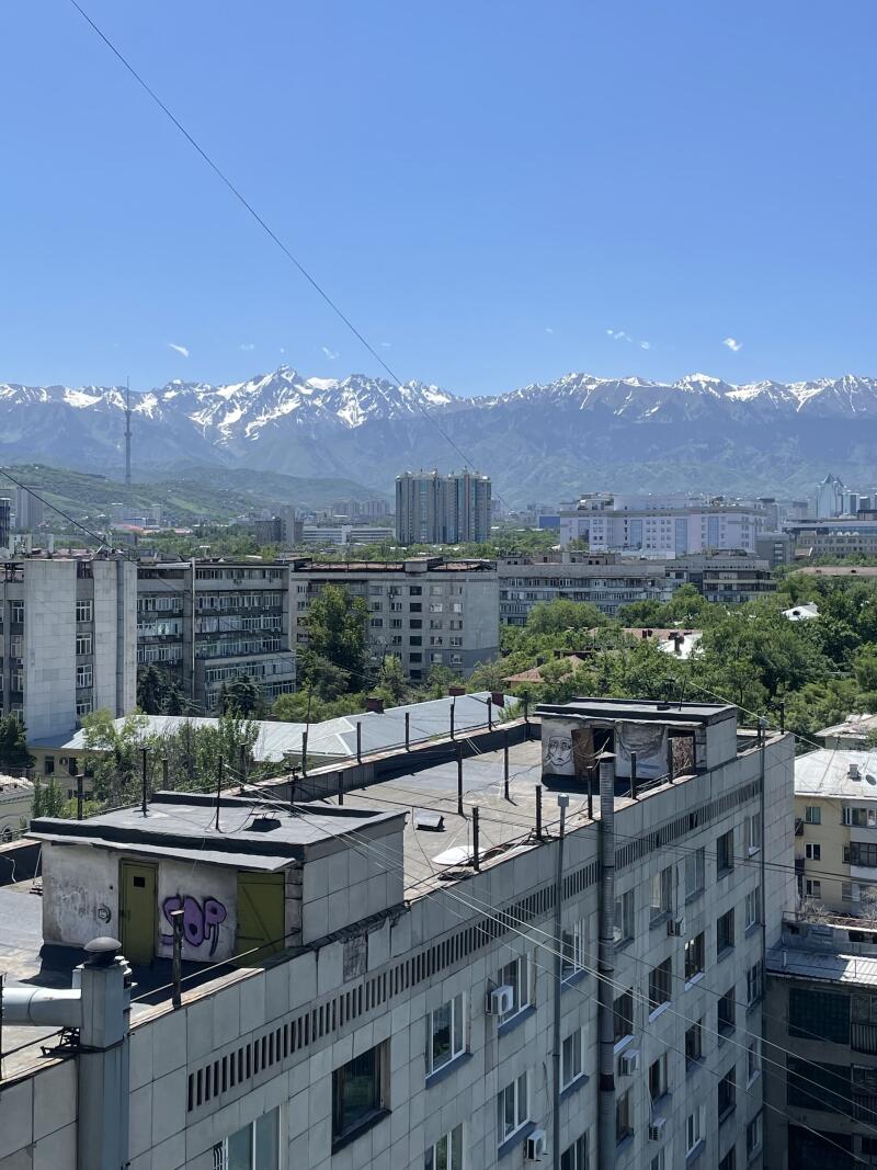 The cityscape features Soviet-era buildings with mountain views under a clear blue sky, some buildings have graffiti.