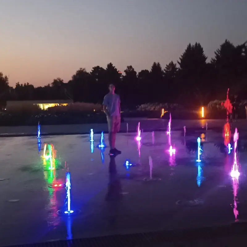 A man stands among colorful, illuminated fountains at dusk.