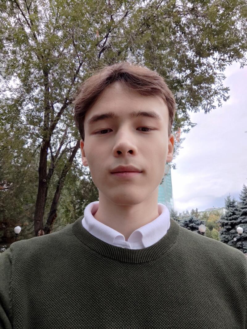 A young man with brown hair is wearing a green sweater over a white collared shirt, standing outside with trees and a building in the background.