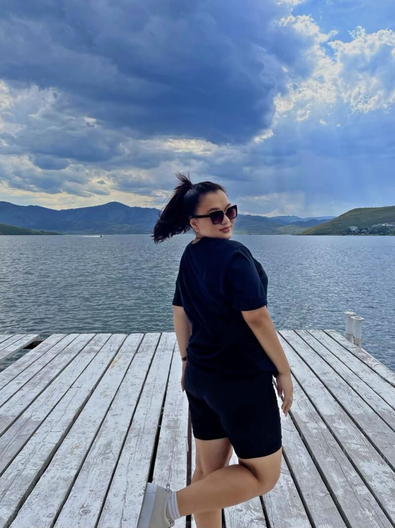 A woman wearing sunglasses, a black T-shirt, and black shorts stands on a white wooden dock, with a body of water and mountains in the background under a cloudy sky.