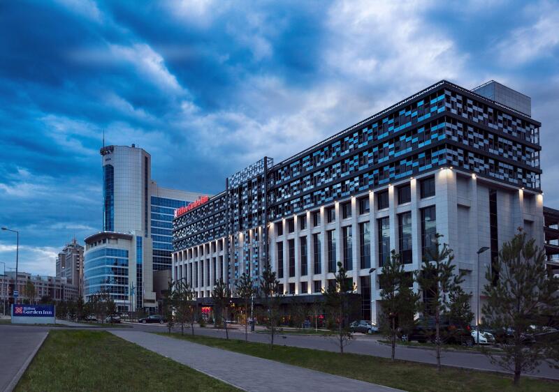 The image shows a modern building with the 'Hilton Garden Inn' sign under a cloudy sky, alongside a street with young trees.