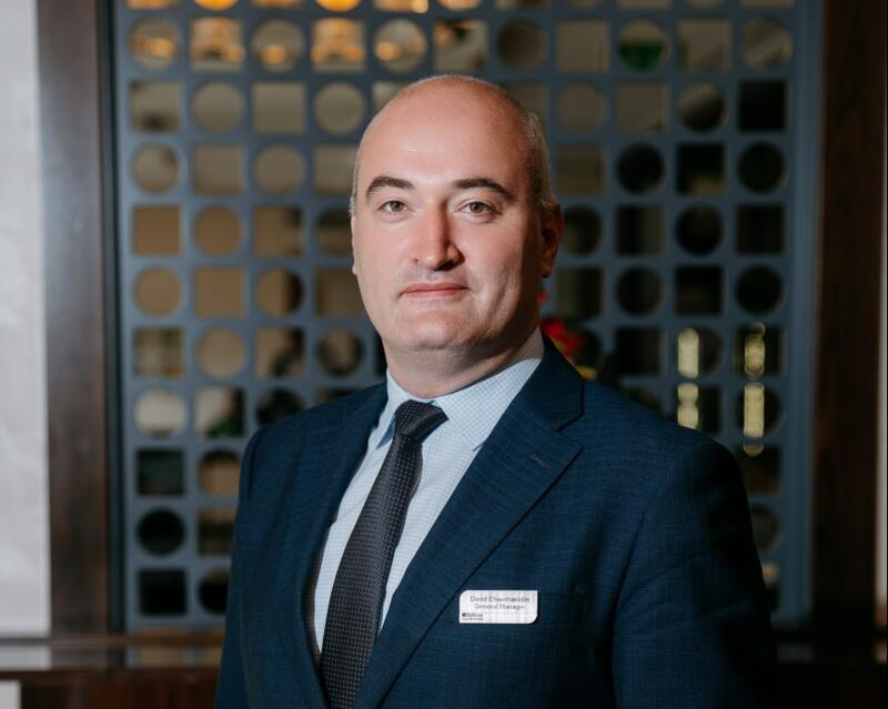 A middle-aged man with a shaved head is wearing a dark blue suit with a dark tie and a name tag that reads "David Chichishvili, General Manager."