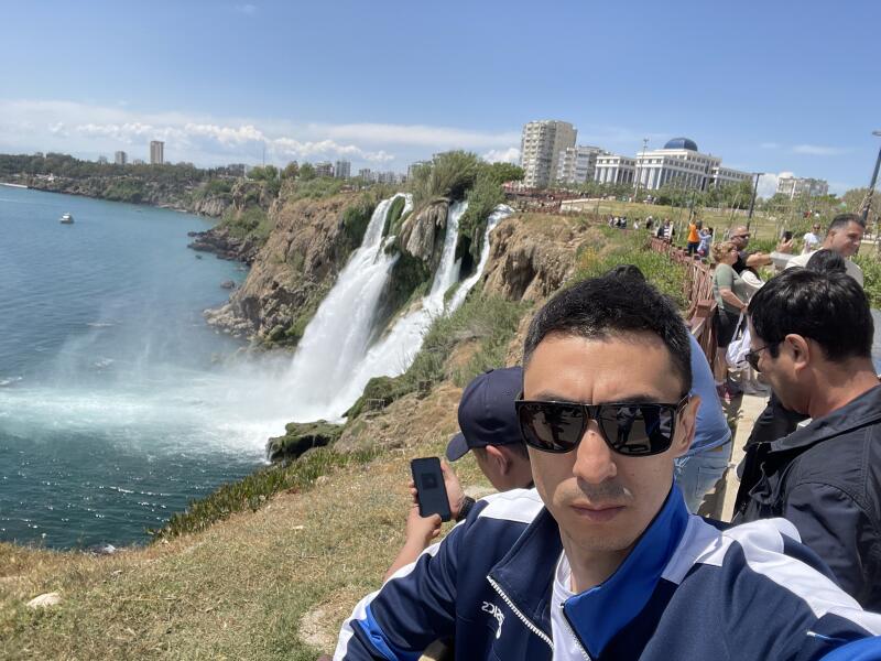 A man in sunglasses and a blue jacket takes a selfie with a waterfall and a coastline in the background on a sunny day.