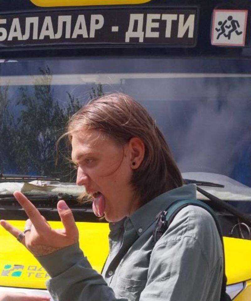 A person with long hair is sticking their tongue out and making a hand gesture in front of a vehicle with the words "БАЛАЛАР - ДЕТИ" and an icon of running children.