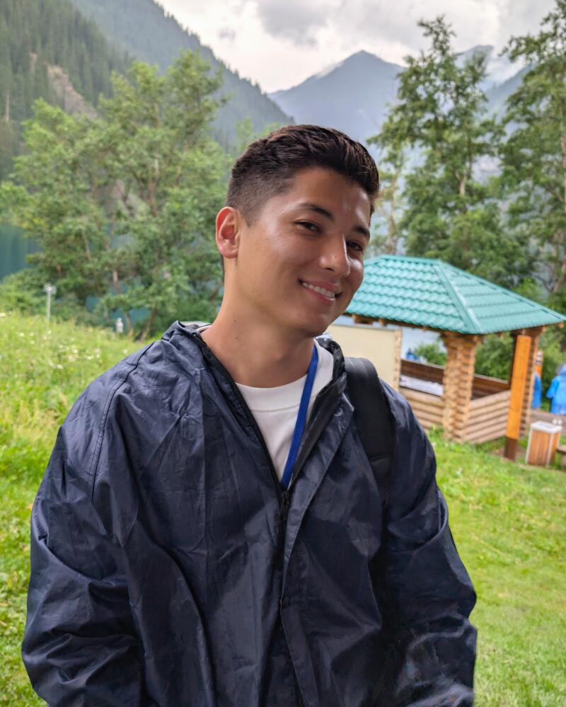 A young man smiles wearing a blue rain jacket and a white shirt with mountains and trees in the background.